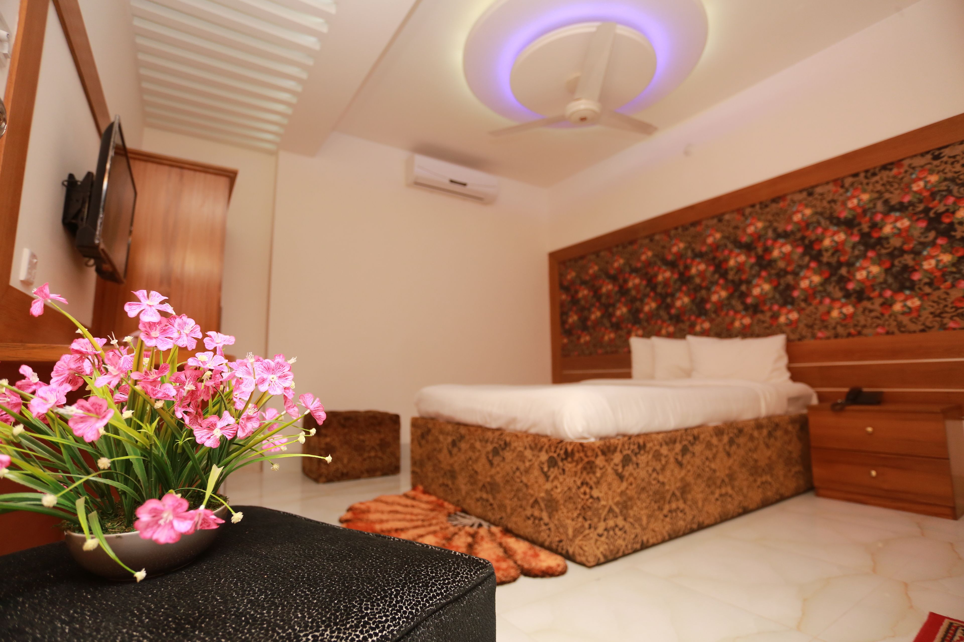 Un dormitorio con cama, mesita de noche, televisor de pared, ventilador de techo y cabecera con motivos florales.