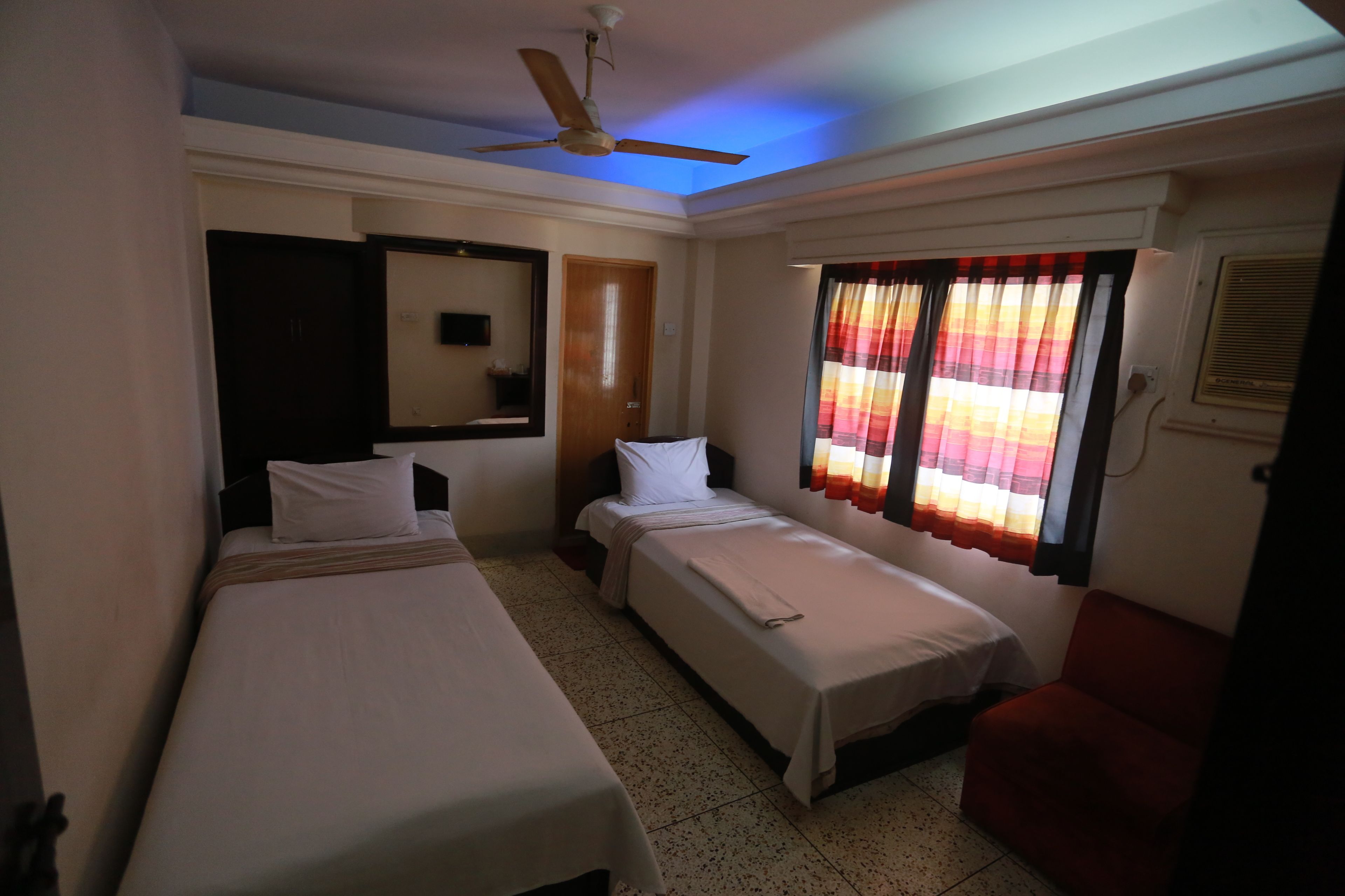 Une chambre d’hôtel avec deux lits simples, un ventilateur de plafond, une fenêtre avec des rideaux et une chaise rouge.