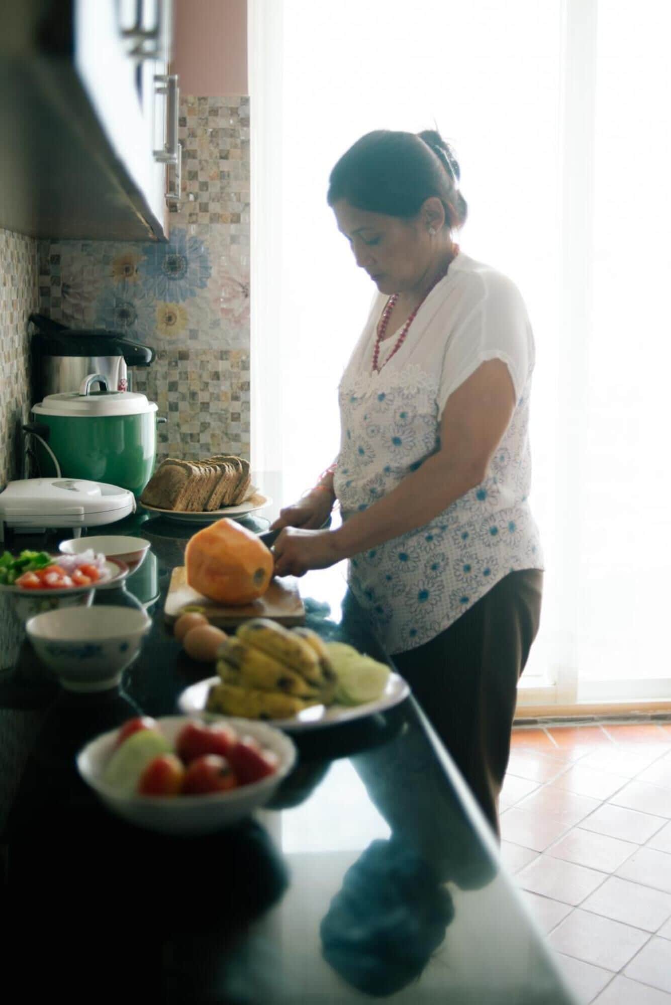A person preparing food in a kitchen with a tiled backsplash, a green slow cooker, and various fruits and vegetables on the counter.