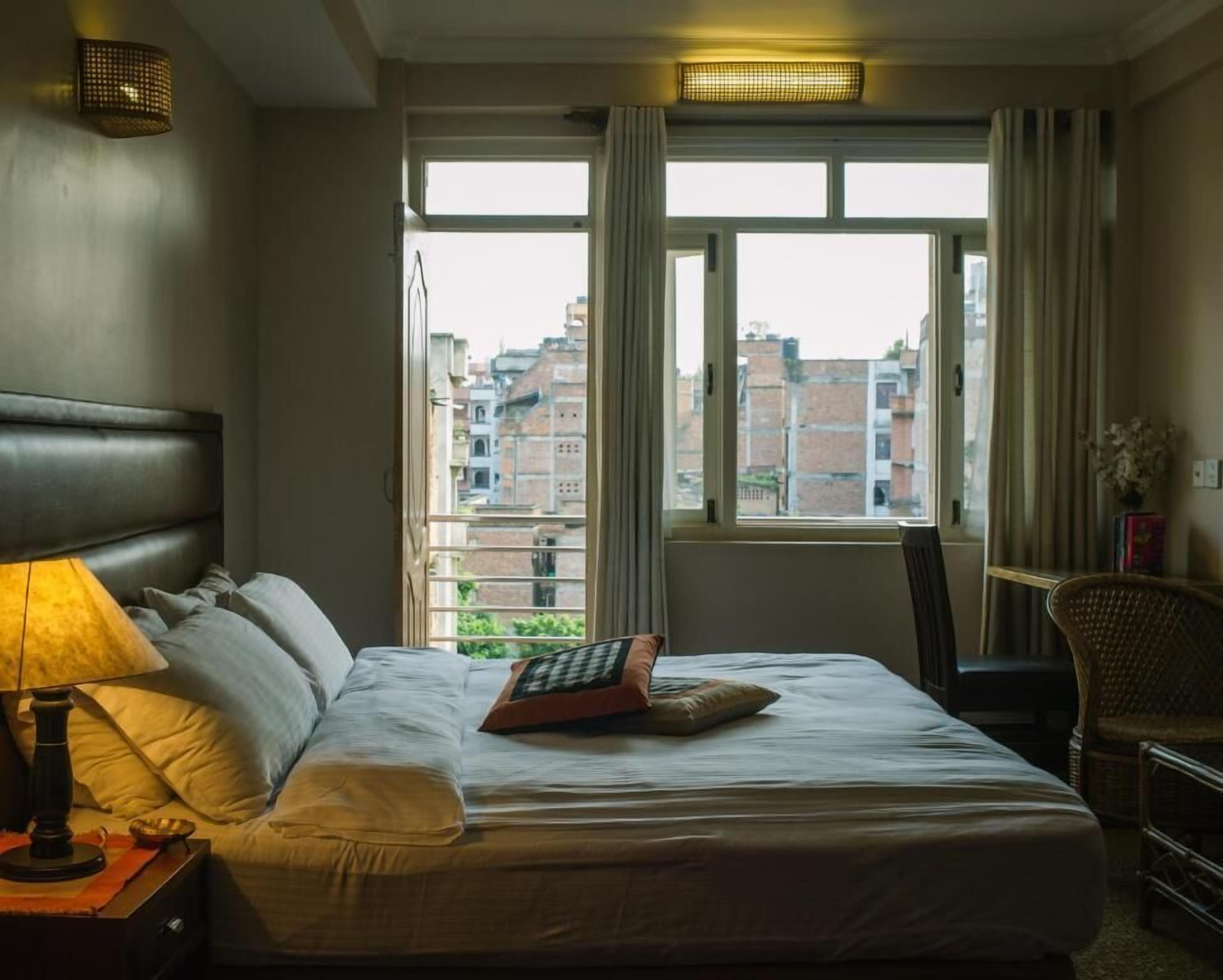 Une chambre d’hôtel avec un lit, une table de chevet, une chaise et une fenêtre donnant sur des immeubles.