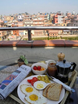 A breakfast tray with eggs, toast, fruit, and a newspaper on a balcony overlooking a cityscape. - Tajaa Pha The Heritage Home (Lalitpur)