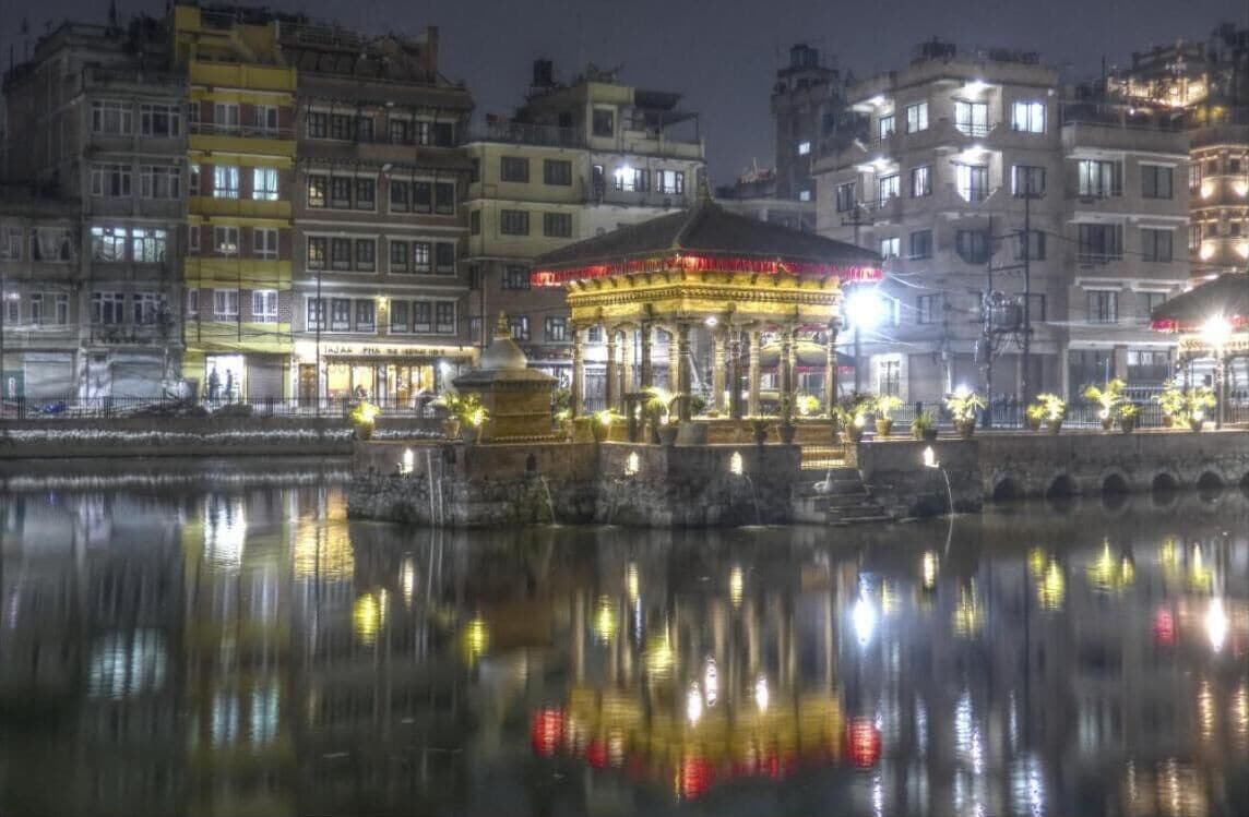 A nighttime view of a riverside area with a well-lit gazebo and buildings reflecting on the water.