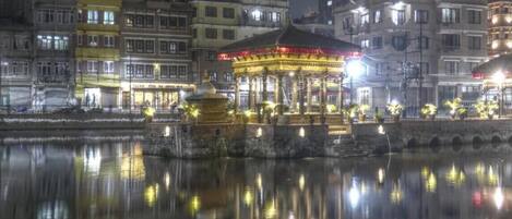 A nighttime view of a riverside area with a well-lit gazebo and buildings reflecting on the water.