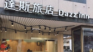 A Dazzinn hotel exterior with a sign in a foreign language, a glass entrance, and outdoor seating with chairs and tables. - Dazz Inn (Taichung)