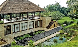 Un bâtiment de style traditionnel, doté d’un toit de chaume, d’un espace extérieur couvert aménagé en coin salon et d’un élément décoratif aquatique agrémenté de nénuphars.