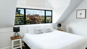 Une chambre avec une grande fenêtre, un lit recouvert de draps blancs, deux tables de chevet avec des lampes et un tableau encadré au mur.