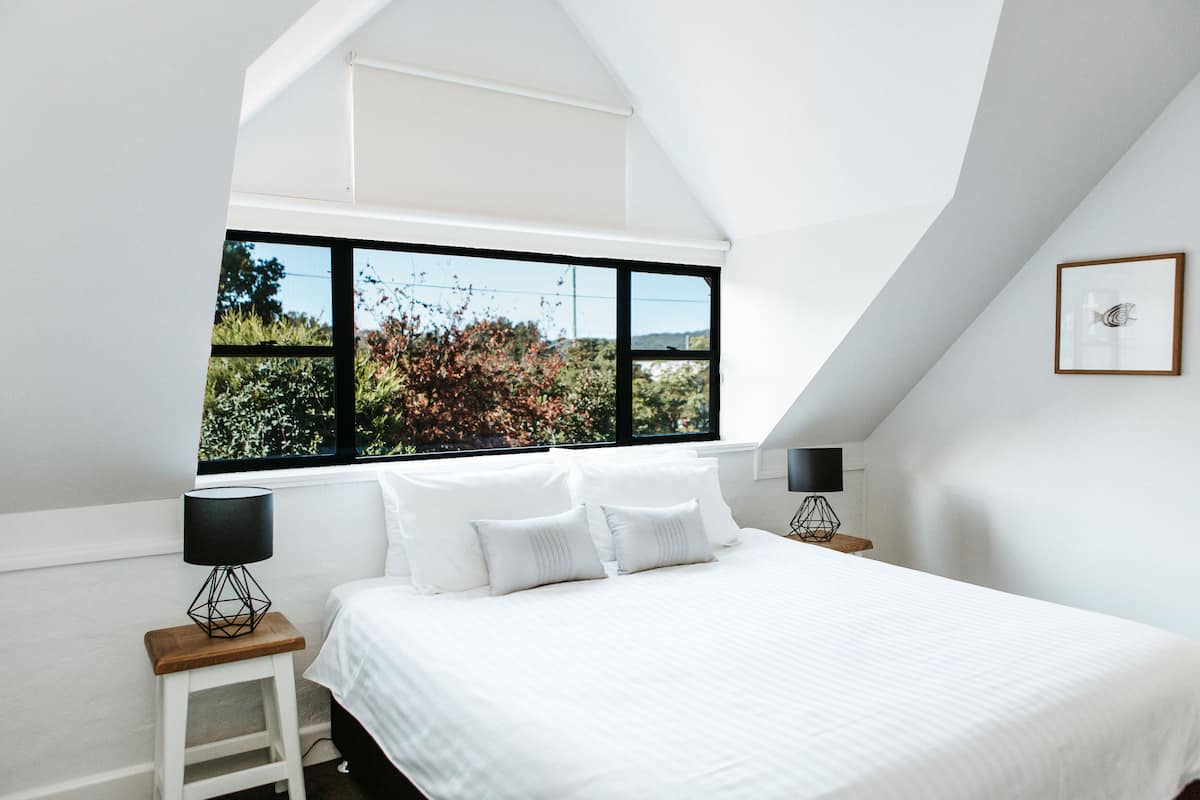 Un dormitorio con un ventanal grande, una cama con ropa de cama blanca, dos mesitas de noche con lámparas y un cuadro en la pared.