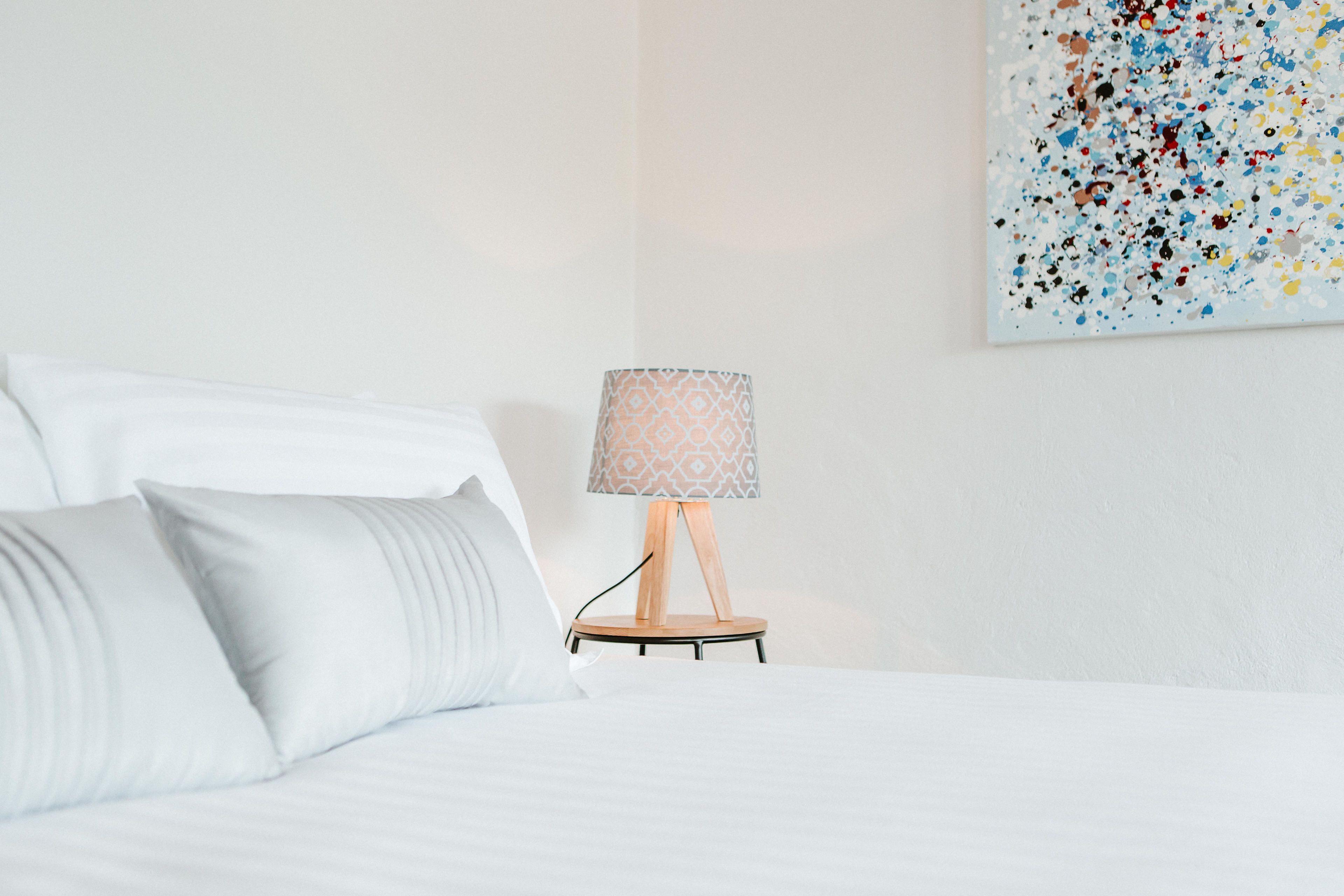A neatly made bed with white linens and pillows, a small wooden bedside table with a patterned lampshade, and a colorful abstract painting on the wall.