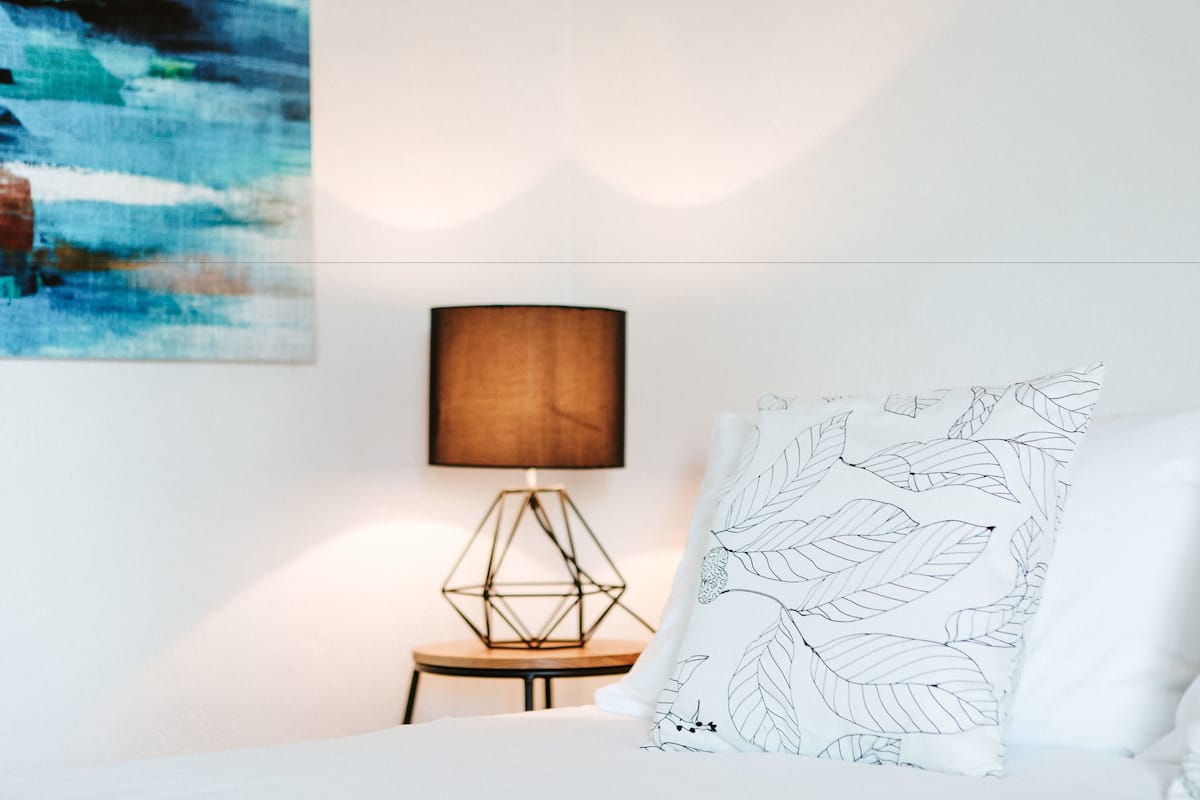 Un dormitorio con una cama, una lámpara geométrica, un cuadro en la pared y un cojín con estampado de hojas.