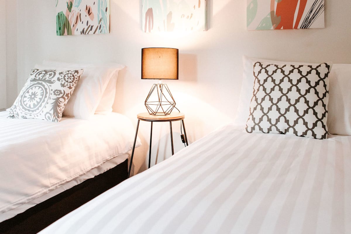 Habitación de hotel con dos camas, una mesita de noche con lámpara y tres obras de arte abstractas en la pared.