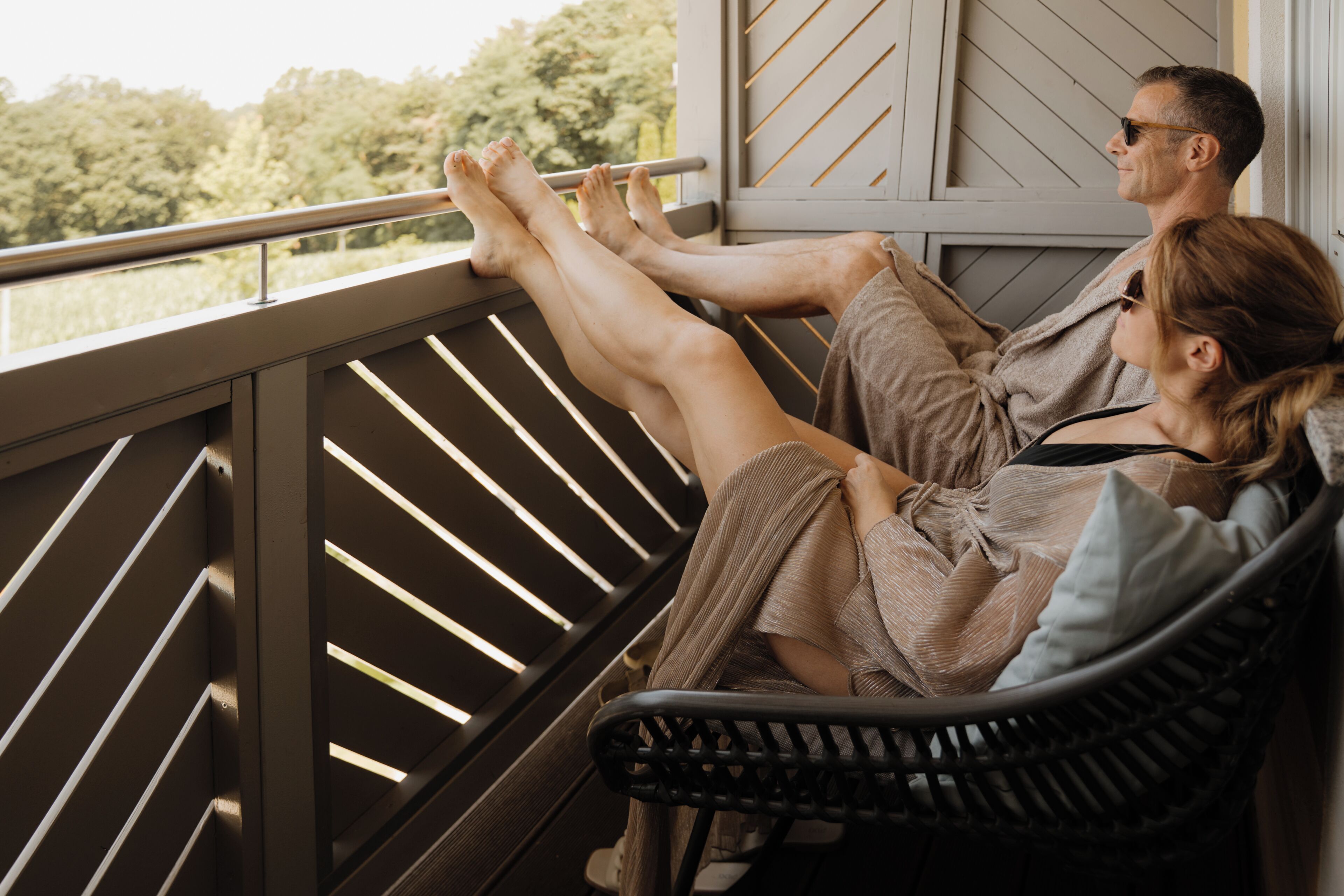 A couple relaxing on a balcony with a view of trees and a railing.