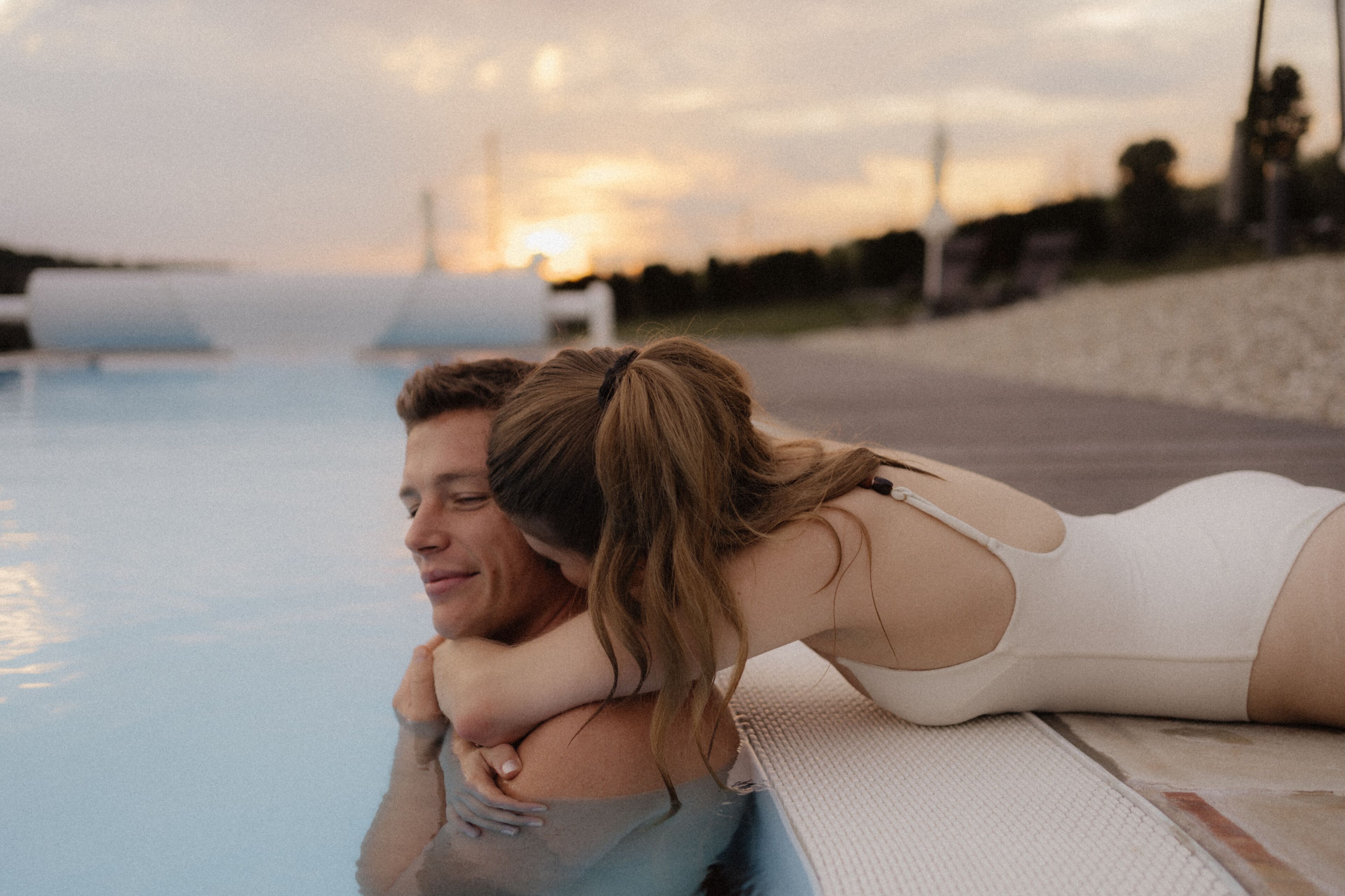 Una pareja se abraza junto a una piscina con un atardecer de fondo.