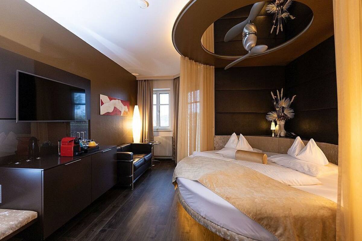 Quarto de hotel moderno com uma cama grande, uma escrivaninha com cadeira, uma TV e um design único para o ventilador de teto.