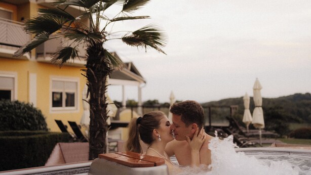 A couple in a hot tub with a palm tree and a yellow building in the background. - Maiers Kuschelhotel deluxe - Adults only (Jennersdorf)
