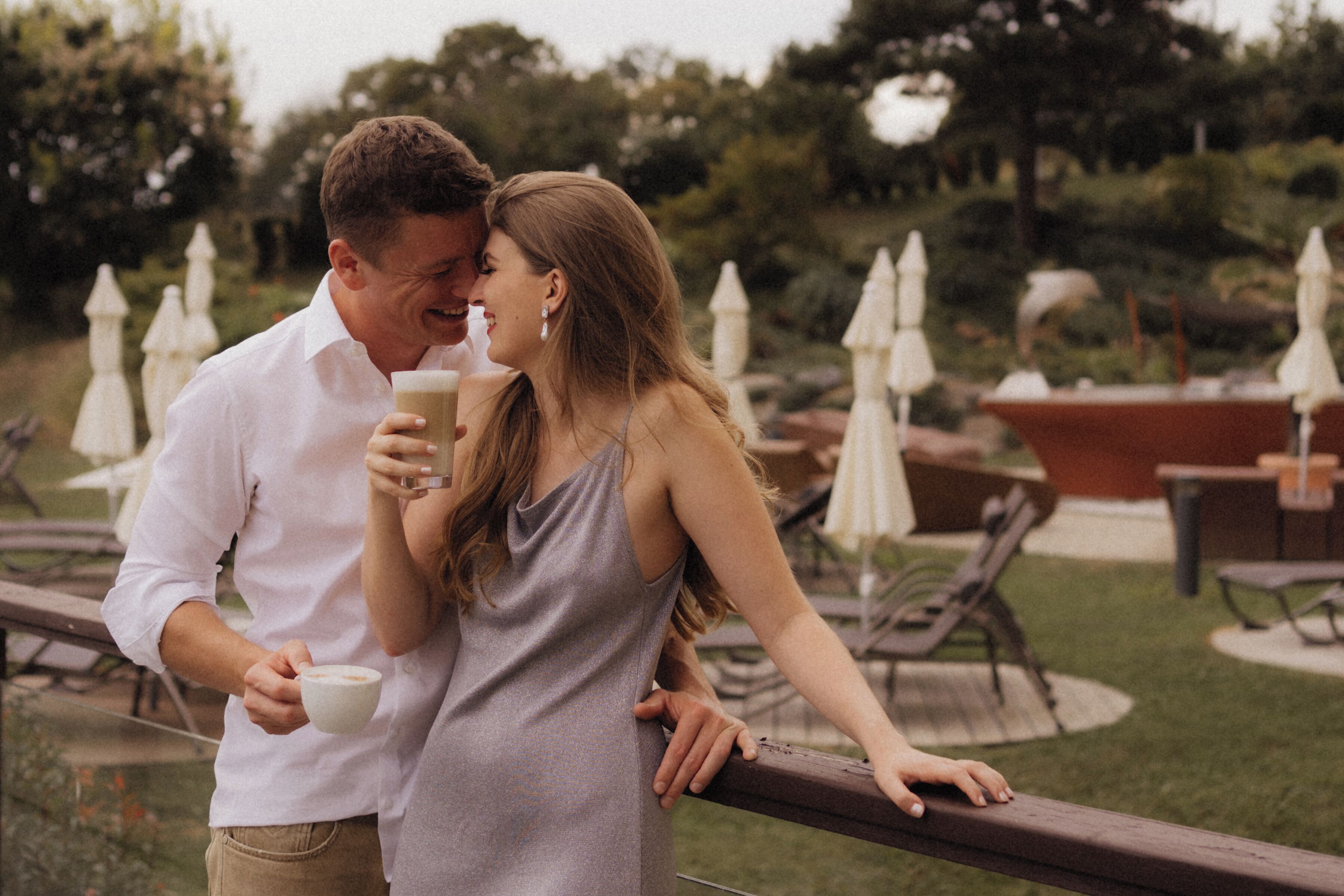 Una pareja sosteniendo tazas de café, de pie junto a una barandilla en un espacio exterior con mesas y sombrillas.