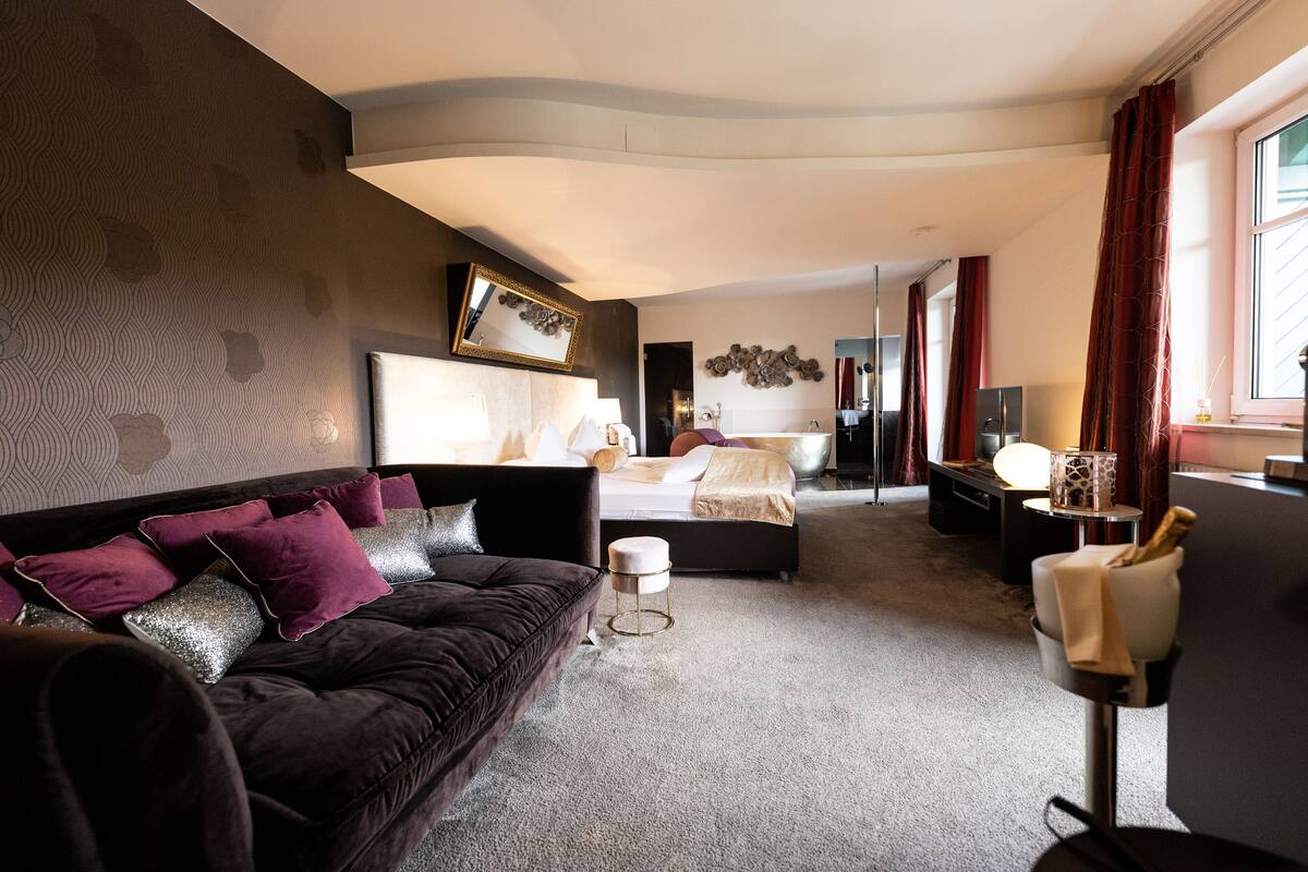 Quarto de hotel moderno com um sofá marrom escuro, uma cama com lençóis brancos, uma mesa redonda pequena e um espelho na parede.