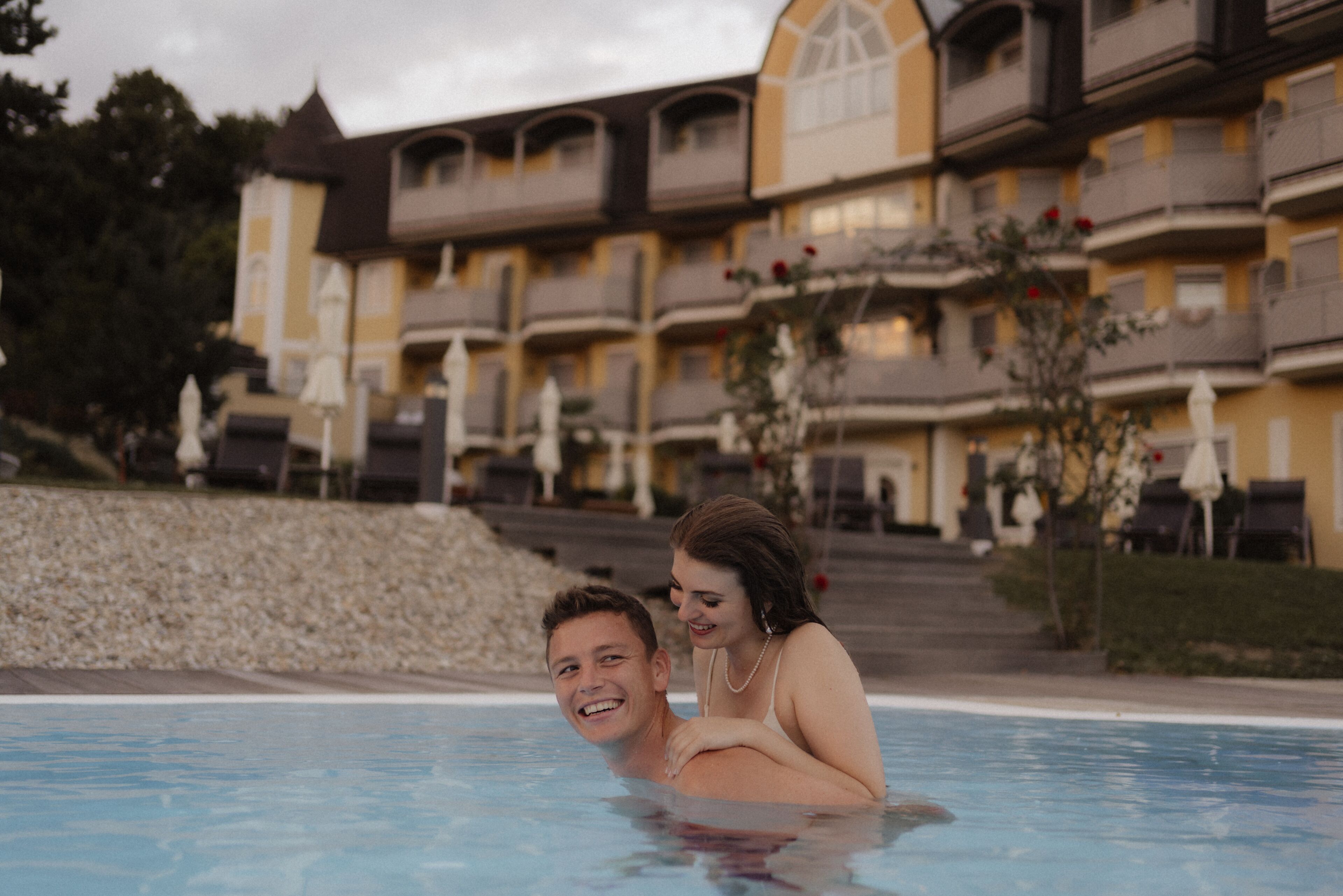 A hotel with a pool area, featuring a building with balconies and a stone wall.