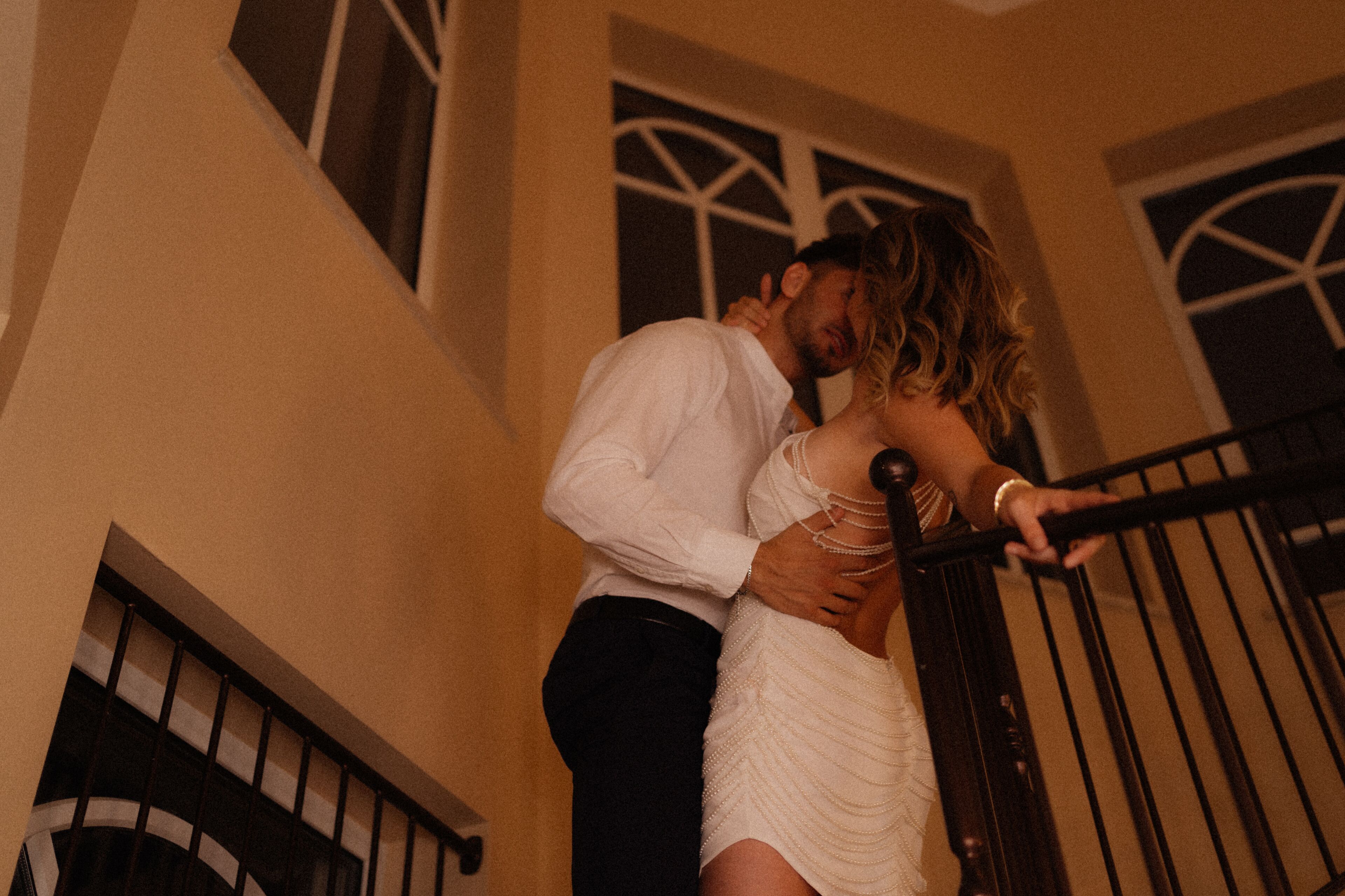 A couple embracing on a staircase with a railing, a window with a decorative frame, and a wall with a textured pattern.