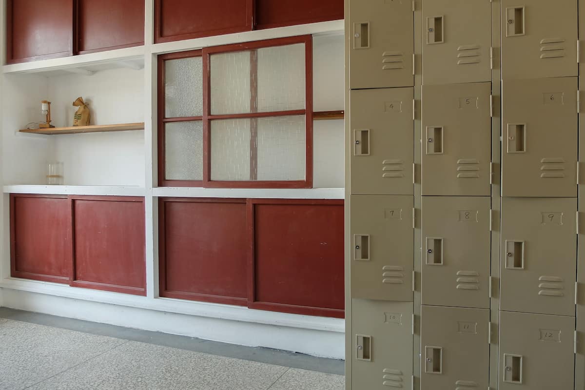 A room with a red and white wall, a window with blinds, and a set of beige lockers with numbers on them.