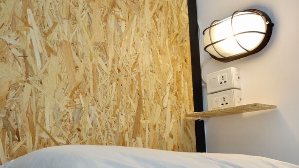 Une chambre avec un mur en bois, un lit avec une literie blanche, un luminaire fixé au mur et une prise de courant.