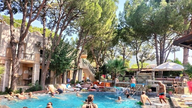 A pool area with multiple people swimming and relaxing, surrounded by trees and a building with balconies. - Atilla's Getaway (Selçuk)