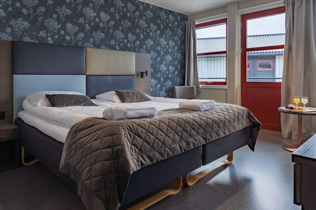 A hotel room with a bed, a chair, a small table with drinks, and a window with curtains.