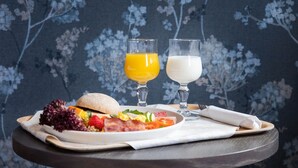 Bandeja de desayuno con un sándwich, ensalada y dos vasos de jugo de naranja y leche, sobre una mesa con un fondo de papel tapiz floral.