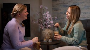 Dos personas sentadas en una mesa con bebidas, un jarrón con flores y un cuadro en la pared.