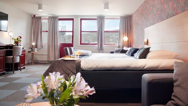 Habitación de hotel con cama, escritorio y silla, vista al exterior y papel tapiz floral en una pared.