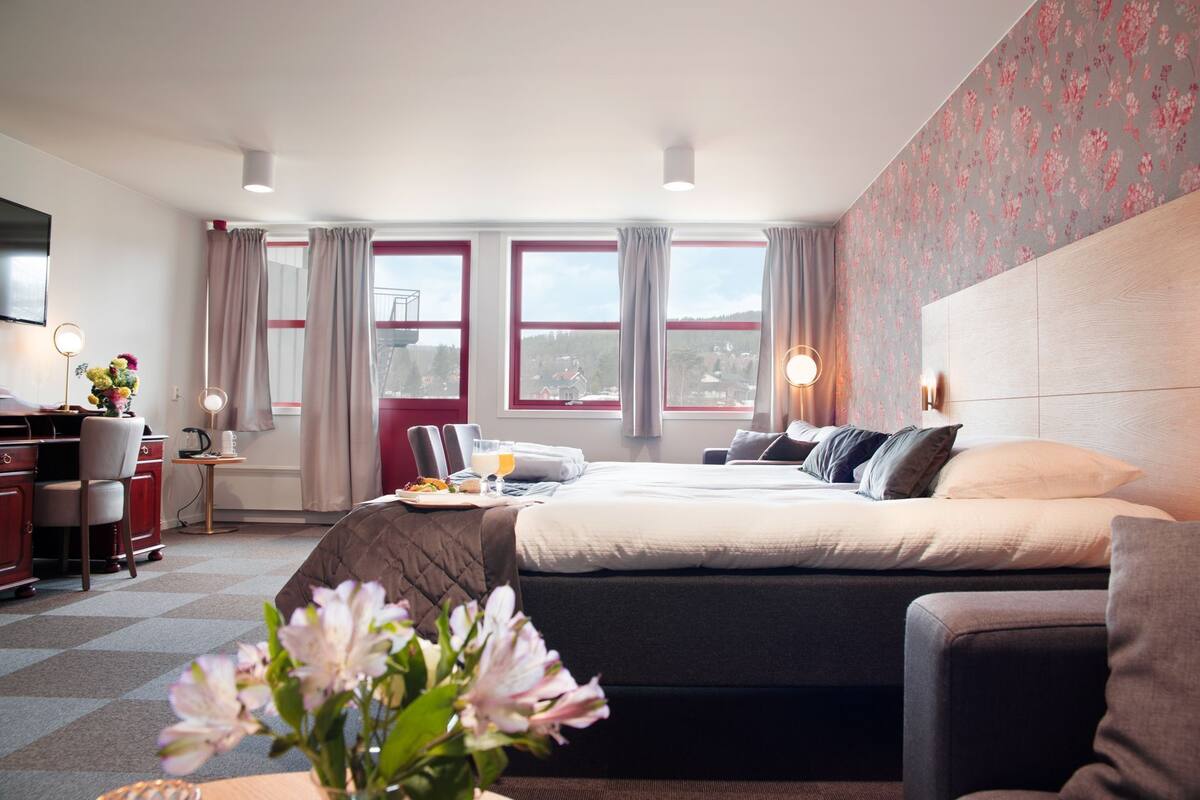 A hotel room with a bed, desk, and chair, a view of the outside, and a floral wallpaper on one wall.