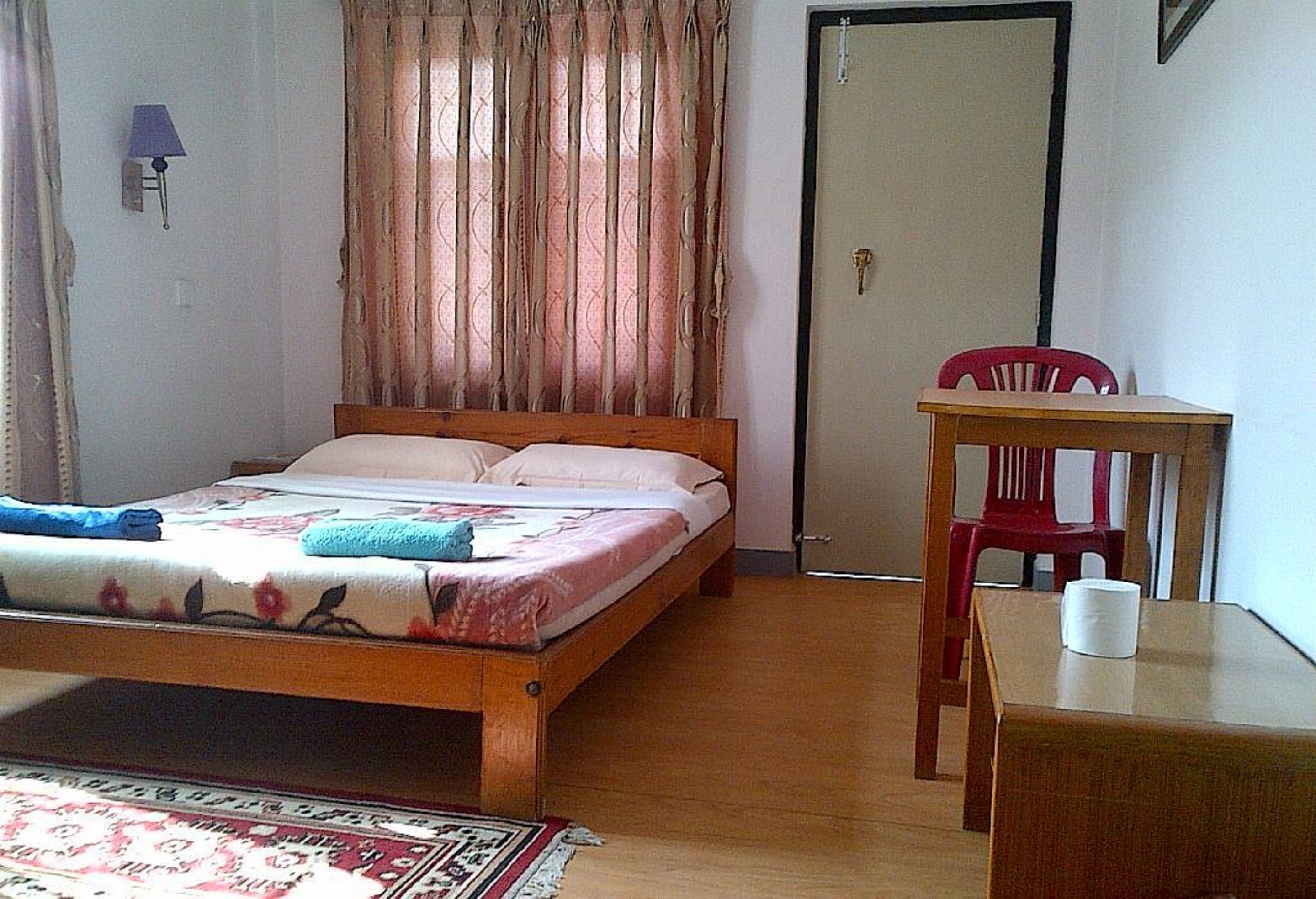 A single bed with a floral bedspread, a wooden nightstand, a red chair, and a small table with a white object on it.