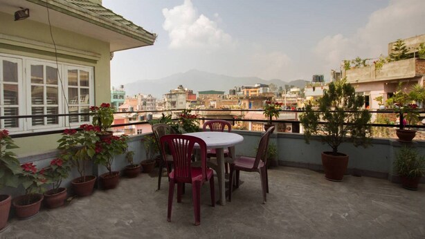 A terrace with a table and chairs, potted plants, and a view of a cityscape with buildings and mountains in the background. - Le Rhododendron Chambres D'hotes (Kathmandu)