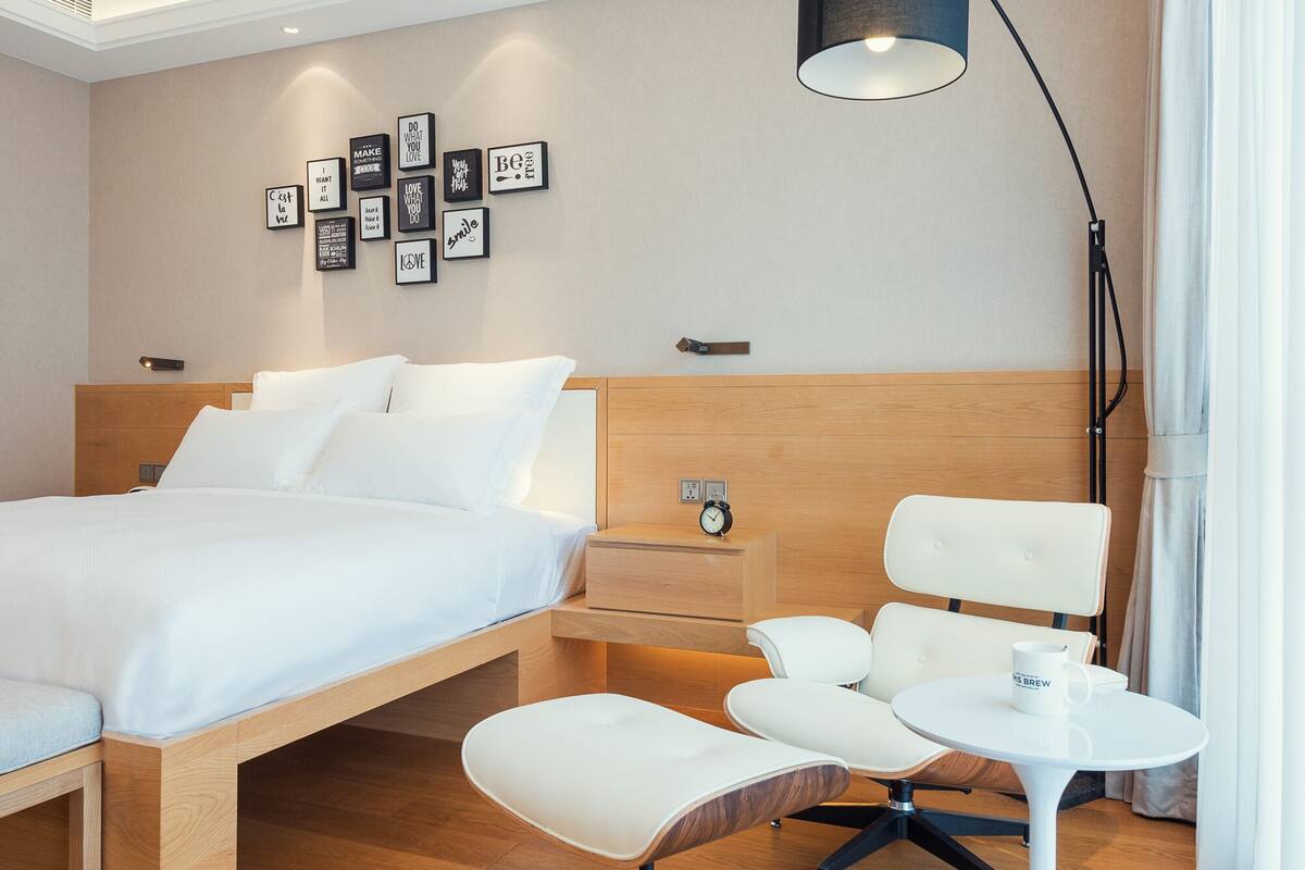 A modern hotel room with a bed, bedside table, clock, and a seating area with a table and chairs.