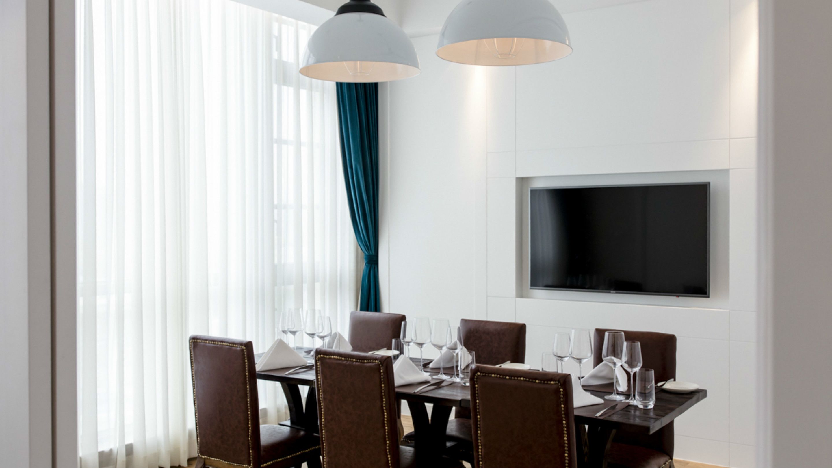 Uma área de jantar com mesa posta para seis pessoas, seis cadeiras, uma TV de tela plana na parede e uma janela com cortinas leves.
