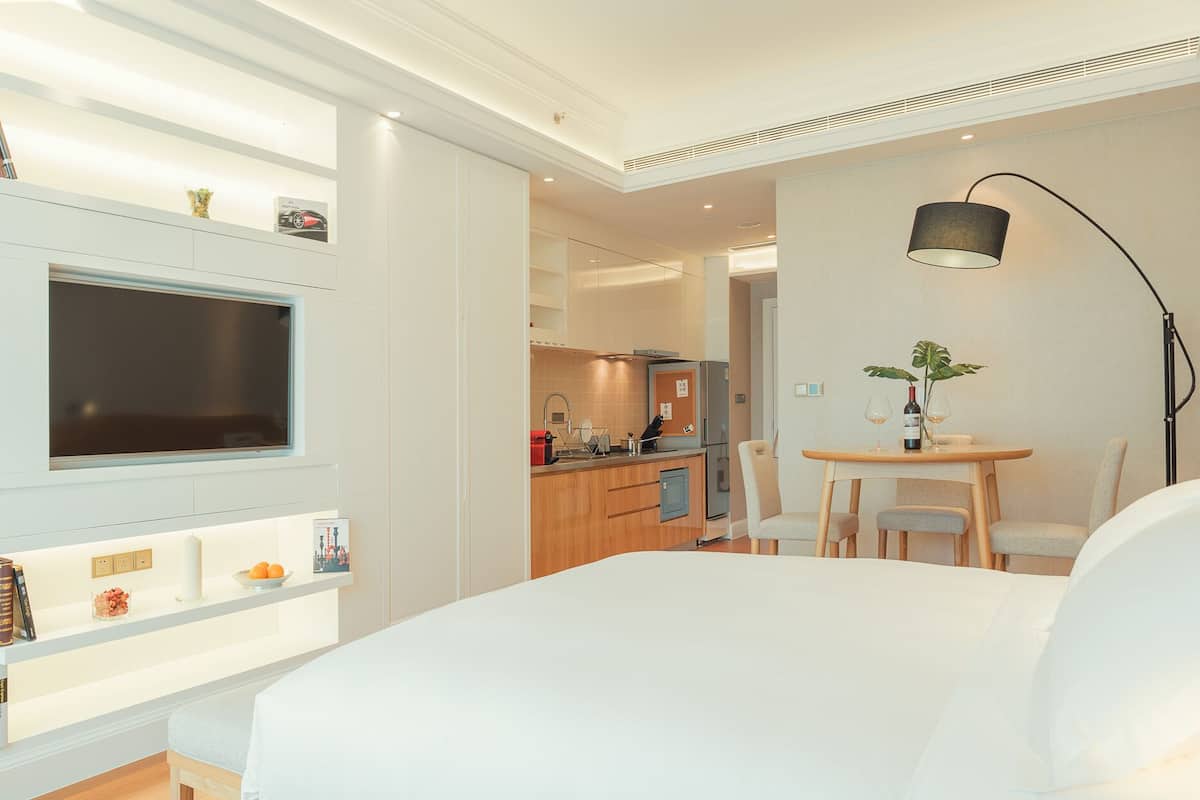 A modern hotel room with a large bed, a TV mounted on a wall, a small dining table with chairs, and a kitchenette in the background.