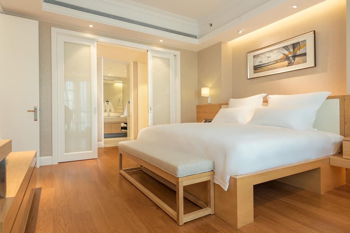 A modern hotel room with a large bed, a bedside table, a nightstand, a mirror, and a painting on the wall.