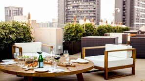 A rooftop dining area with round tables, wicker chairs, and a city view.