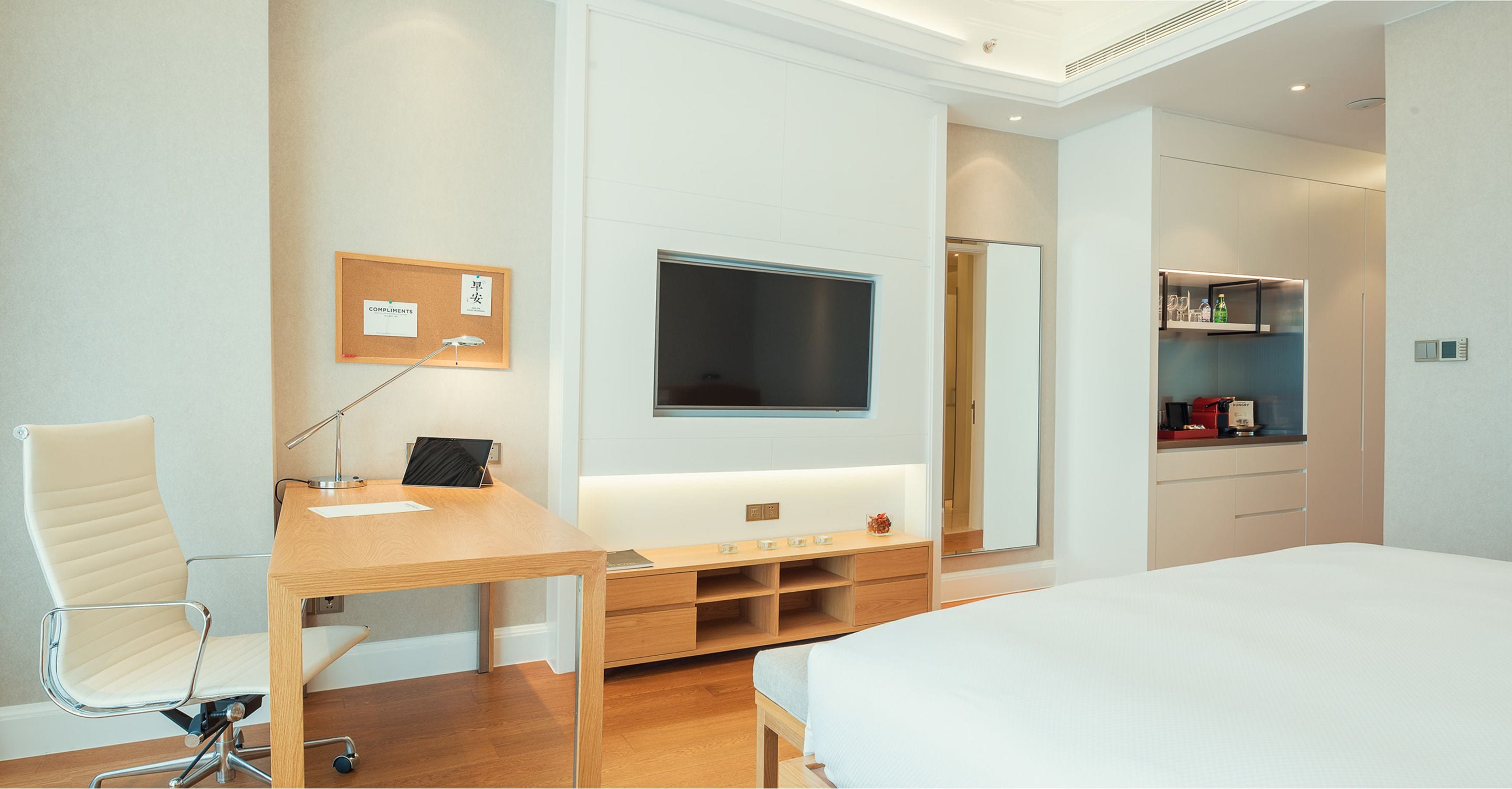 Une chambre d’hôtel moderne avec un bureau en bois, une chaise blanche, un lit, un téléviseur fixé au mur et un petit coin cuisine.