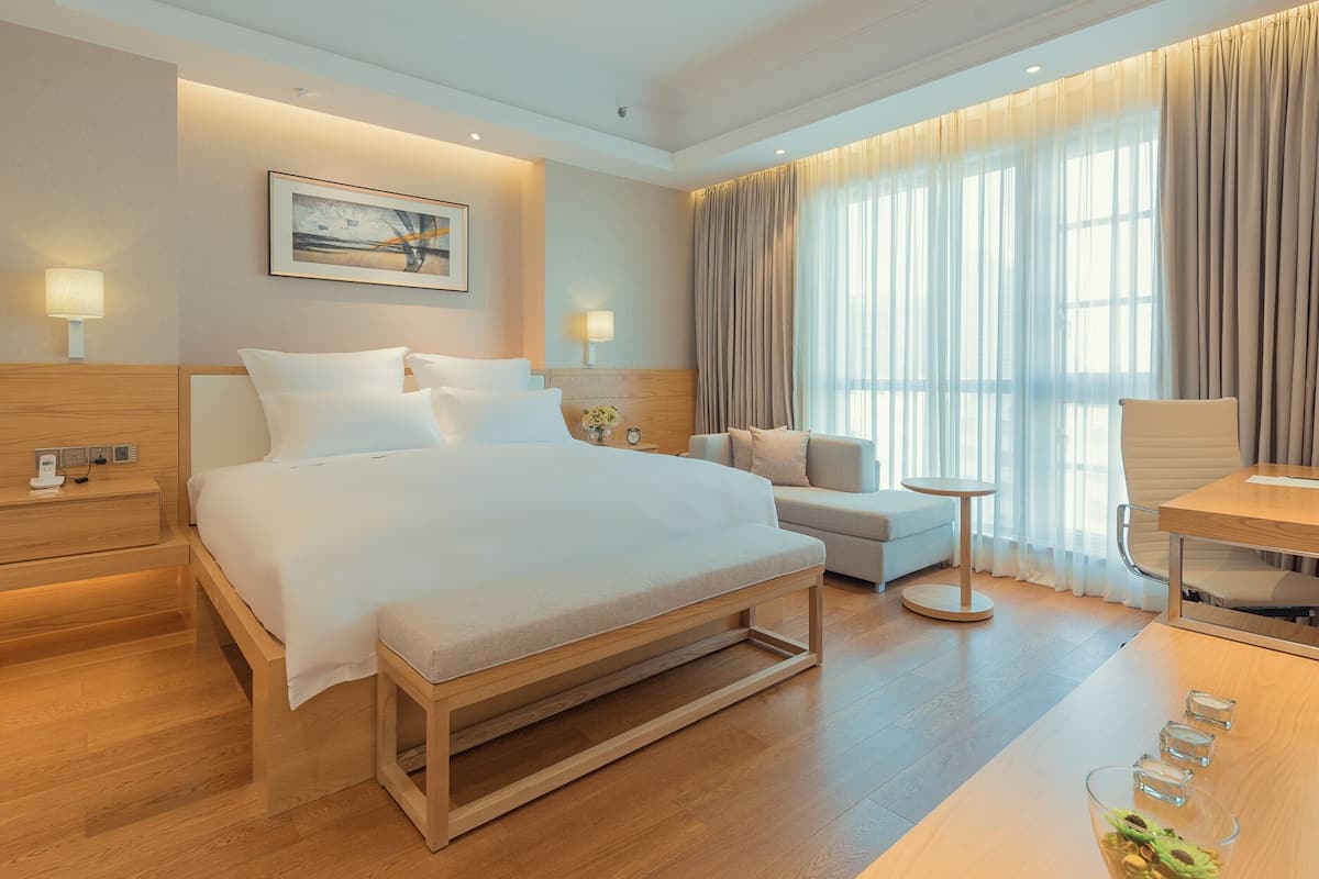 A modern hotel room with a large bed, a bedside table, a small sofa, a round table, and a desk.