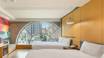 A hotel room with a large window, two beds, a desk, and a view of the cityscape.