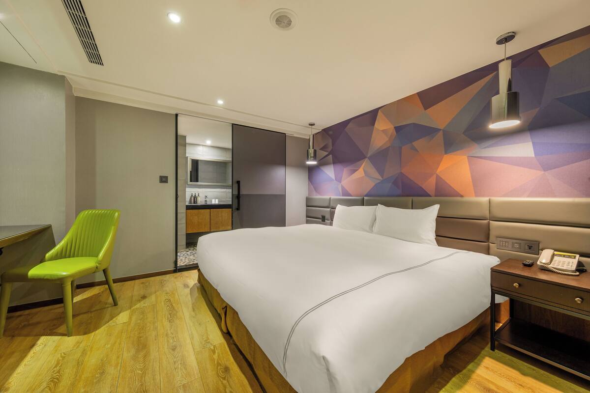 A modern hotel room with a large bed, a desk, a chair, and a geometric-patterned wall.