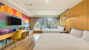 Quarto de hotel moderno com uma cama grande, uma escrivaninha com cadeira, televisão e vista para a cidade através de uma janela ampla.
