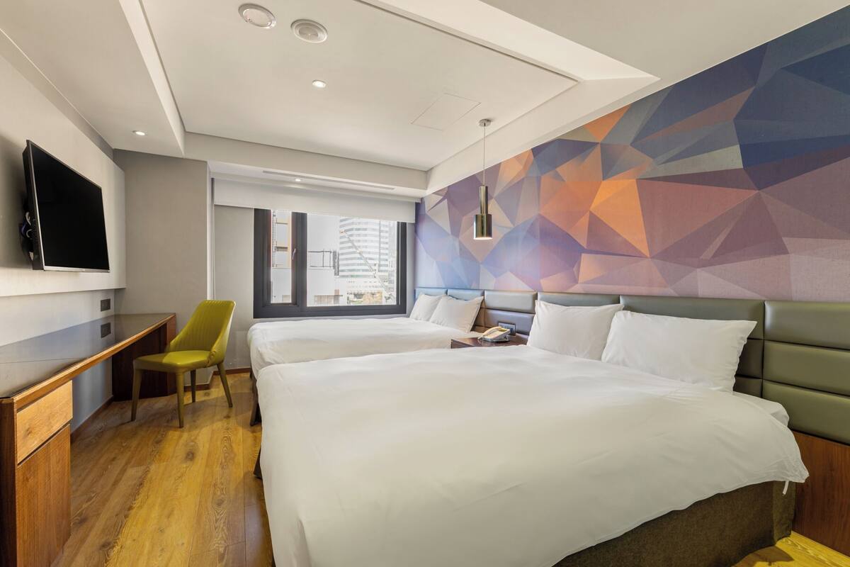 A hotel room with two beds, a desk, a chair, a TV, and a geometric-patterned wall.