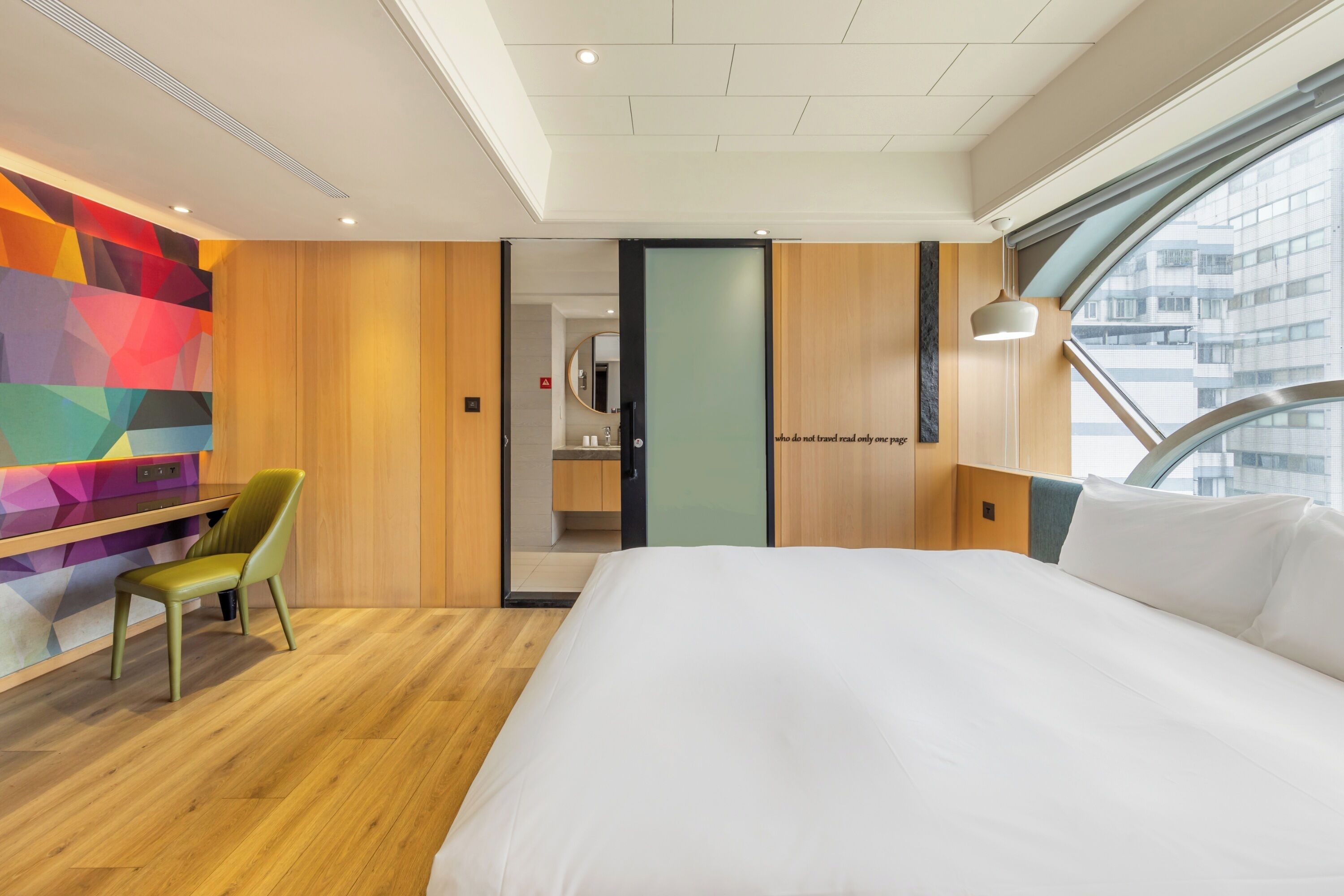 Une chambre d’hôtel moderne avec un grand lit, un plancher en bois, un mur aux motifs géométriques et colorés, et une vue sur la ville par une grande fenêtre.