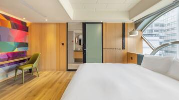 Une chambre d’hôtel moderne avec un grand lit, un plancher en bois, un mur aux motifs géométriques et colorés, et une vue sur la ville par une grande fenêtre.