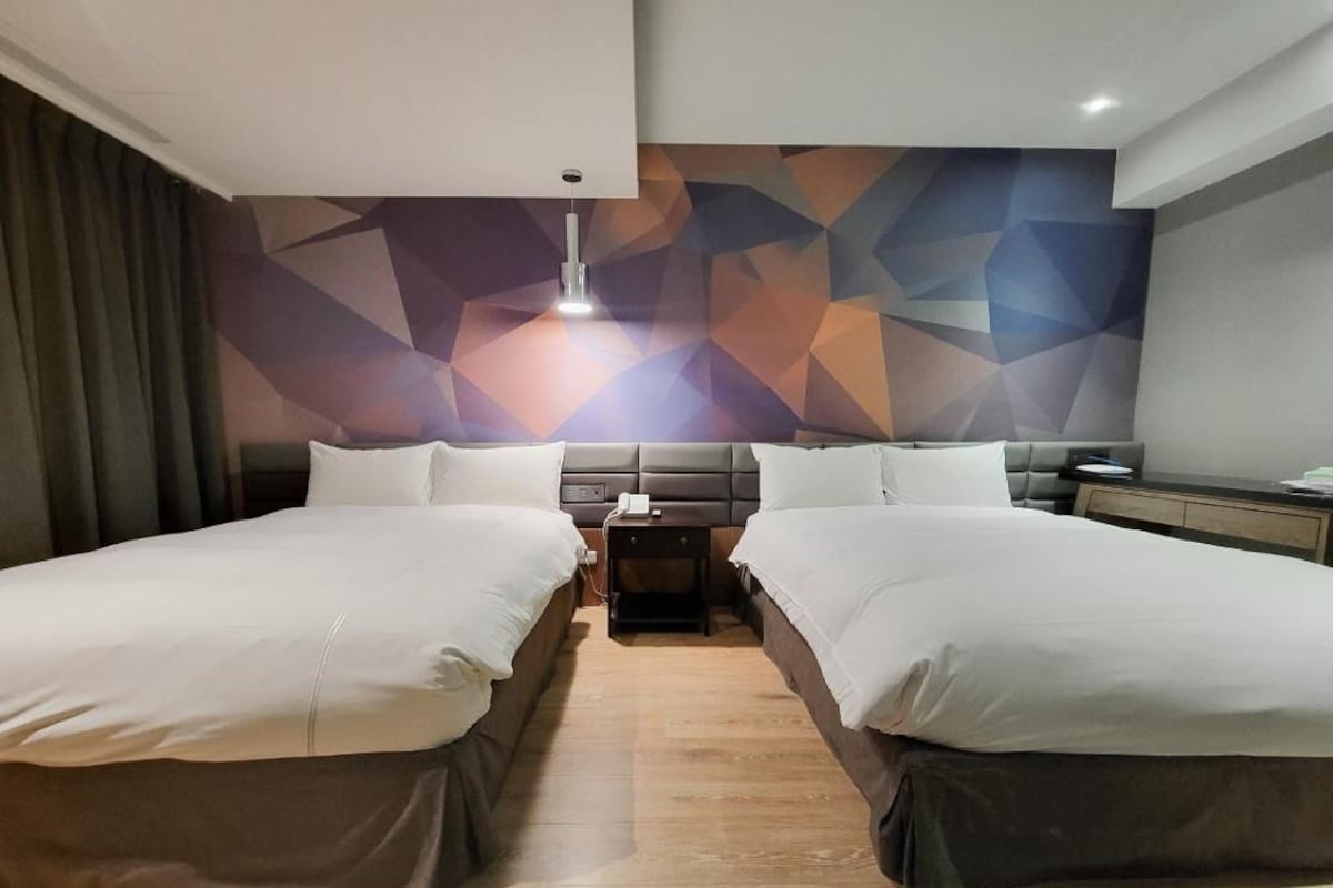 A modern hotel room with two beds, a geometric wall design, a bedside table, and a nightstand.