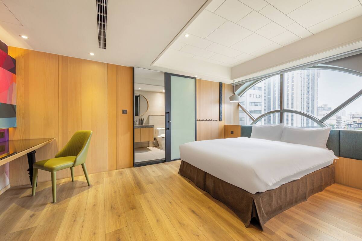 A modern hotel room with a large bed, a desk, and a chair. There is a view of the city through a large window.