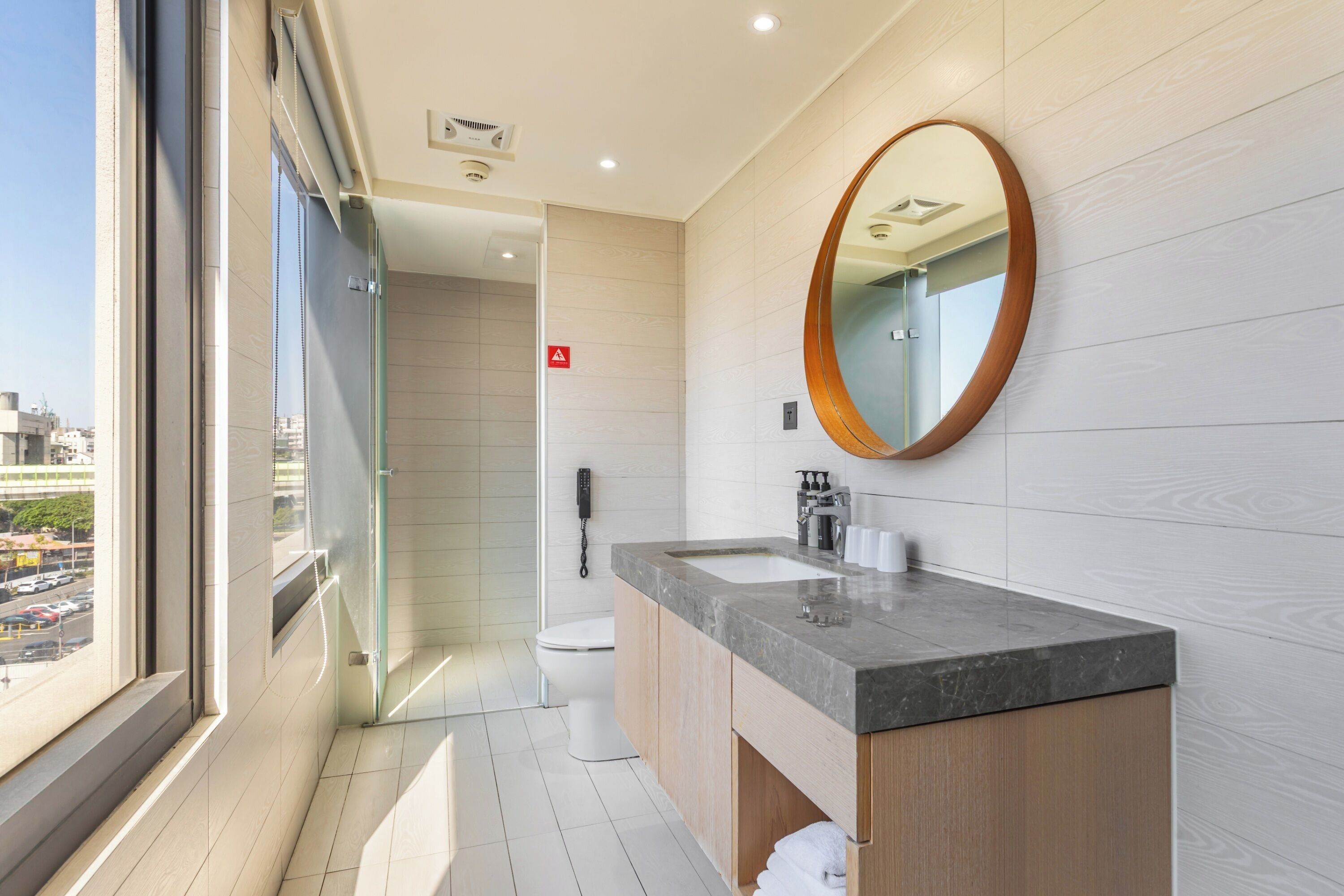 A modern bathroom with a large window, a round mirror, a sink, and a toilet.