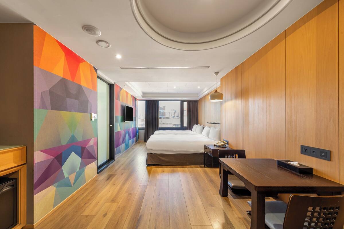 A hotel room with a bed, bedside table, desk, and a colorful geometric wall.