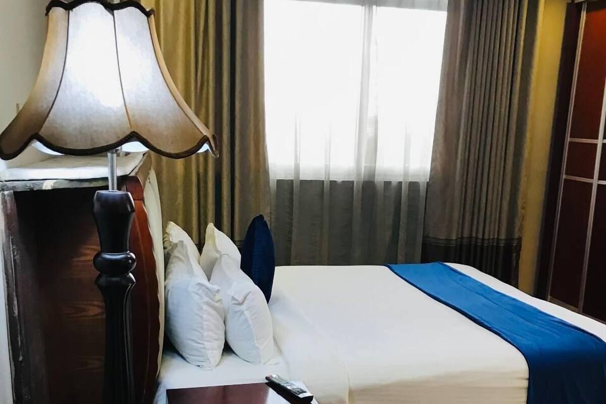 Habitación de hotel con cama, mesita de noche, lámpara, ventana con cortinas y un par de pantuflas en el suelo.